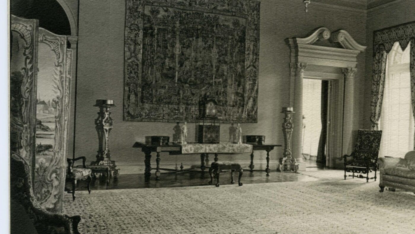 "Filoli Reception Room south wall with large tapestry hanging at center.  Partial view of folding screen on left and arm chairs on each side.  Tall ornate gilt stands on both sides of tapestry. Long rectangular table under tapestry with ornate clock in center and two figurines and two decorative boxes on each side.  Tapestry on table and small needlepoint bench in front of table.  Upholstered side chair with wooden arms in front of window in southwest corner and sconce on west wall.  Floral damask sofa along west wall.  Lower portion of chandelier at top of photo. Large tapestry rug centered on floor with no furniture in center of room. Believe to be early Roth period just after they moved in."