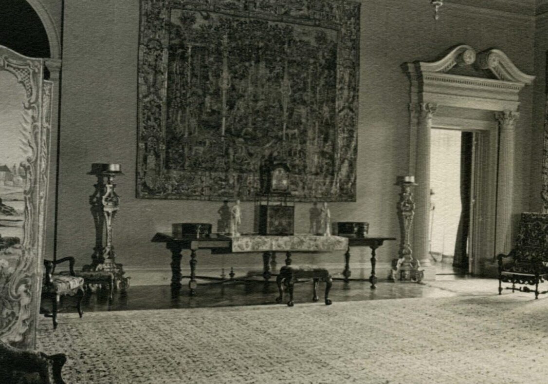 "Filoli Reception Room south wall with large tapestry hanging at center.  Partial view of folding screen on left and arm chairs on each side.  Tall ornate gilt stands on both sides of tapestry. Long rectangular table under tapestry with ornate clock in center and two figurines and two decorative boxes on each side.  Tapestry on table and small needlepoint bench in front of table.  Upholstered side chair with wooden arms in front of window in southwest corner and sconce on west wall.  Floral damask sofa along west wall.  Lower portion of chandelier at top of photo. Large tapestry rug centered on floor with no furniture in center of room. Believe to be early Roth period just after they moved in."