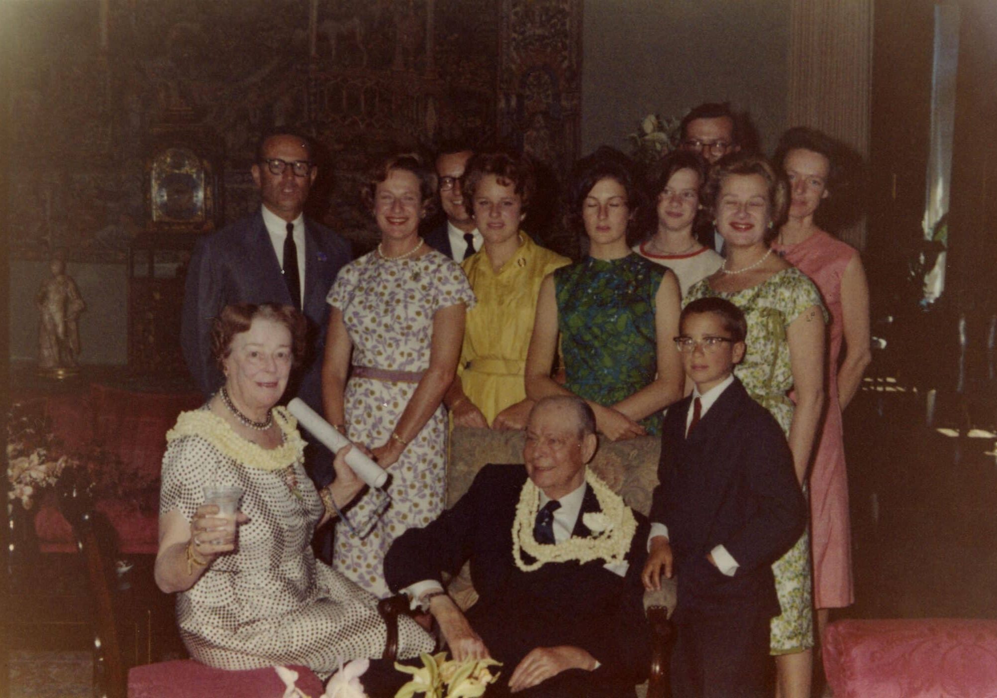 "Birthday party for William Phillip Roth (Bill) family shot in the Reception Room. From left to right, front row: Lurline Berenice Matson Roth seated in a light colored graphic dress, with Bill seated in center, wearing a dark suit and tie, and young James Coonan (Jim)? in a suit; second row: James Coonan (Jim) in a blue jacket and tie, next to his wife Lurline Matson Roth Coonan in a white gown with purple and yellow floral pattern, Kathy Roth? in a yellow belted shirt dress, Lurline Coonan (Lolly) in a bright green and dark blue patterned dress, and Berenice Matson Roth Grant in a light colored floral dress; third row: Unidentified Man in a jacket and tie, next to an Unidentified granddaughter in a light dress with red trim, and Joan Roth? in a dark pink dress; William Matson Roth (Bill) obscured in back in a jacket and tie. Both Lurline and Bill both wear yellow flower leis around their necks, 10 family members stand around Bill seated in pale blue floral decorated library chair. The lower corner of the south wall tapestry is visible in background, along with a standing figure statue on the tabletop behind them, along with an elaborate clock and stand."
